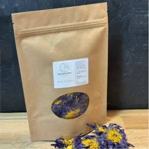 Brown kraft bag labeled Pachamama cacao, with a round window showing purple and yellow dried lotus petals inside; sits on a wooden surface with some petals spilled nearby.