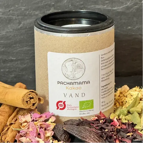 Cylindrical container with a black lid and kraft-label reading 'Pachamama Kakao Vand', surrounded by dried flowers and cinnamon sticks.