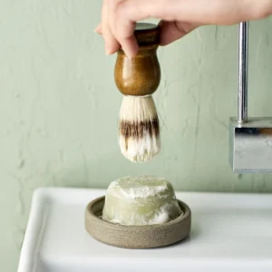 Close-up of a hand pressing a wooden-handled shaving brush into its stand against a pale green wall.