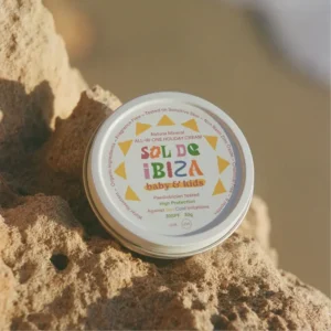 Round tin of Sol de Ibiza baby & kids sunscreen (SPF 30) resting on sandy rock at the beach, warm sunlight on the label.