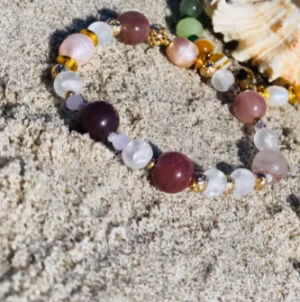 Two beaded bracelets with multicolored stones lie on light sand beside a seashell at the beach.