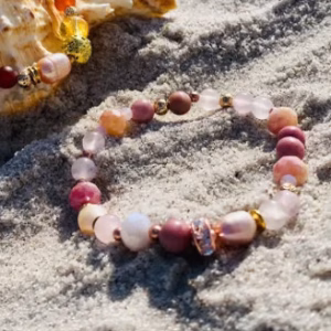 Conch shell and colorful beaded bracelets resting on sandy beach with the sea in the background.