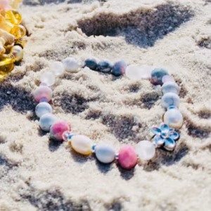 Colorful shell with a beaded bracelet on sunlit beach sand, with the sea in the background.