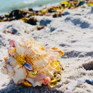Shell and pastel bead bracelet on sandy beach near the waterline, sunlight highlighting the shells and jewelry.