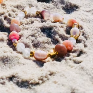 Beige sea shell with colorful beaded bracelet half-buried in sand on a sunny beach; ocean waves in the background.
