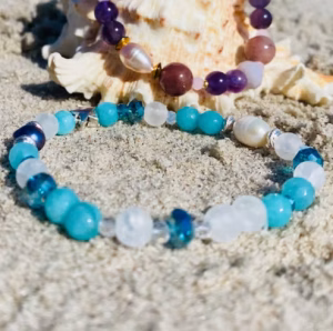 Beaded bracelets in purple, pink, and gold draped over a seashell on sandy beach with ocean in the background (beach jewelry display).