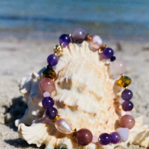 Beaded bracelets draped over a large seashell on a sandy beach with the ocean in the background, purple, pink, and turquoise beads visible.