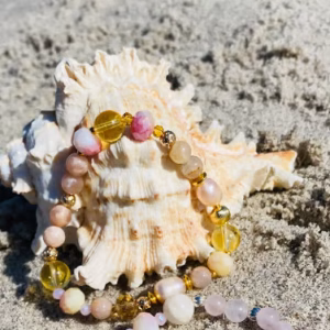 Pastel beaded bracelet and necklace arranged around a seashell on sandy beach, with pink, yellow, and clear beads.