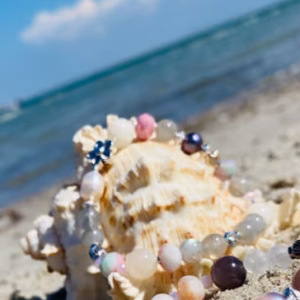 Pastel bead bracelet draped around a seashell on a sandy beach with the sea in the background, star charm visible.
