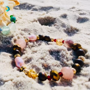 Colorful beaded bracelet lays in the sand beside a large seashell on a sunny beach with the ocean in the background.