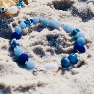 Blue and white beaded bracelet resting on sandy beach beside a large seashell, ocean in the background on a sunny day.