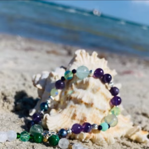 Colorful beaded bracelet resting on sandy beach with a seashell nearby.